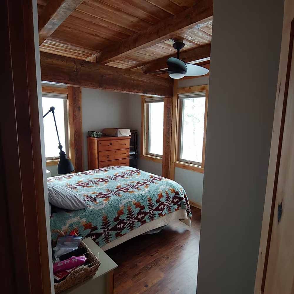 Cozy wooden bedroom with quilted bed, dresser, windows, and exposed beam ceiling