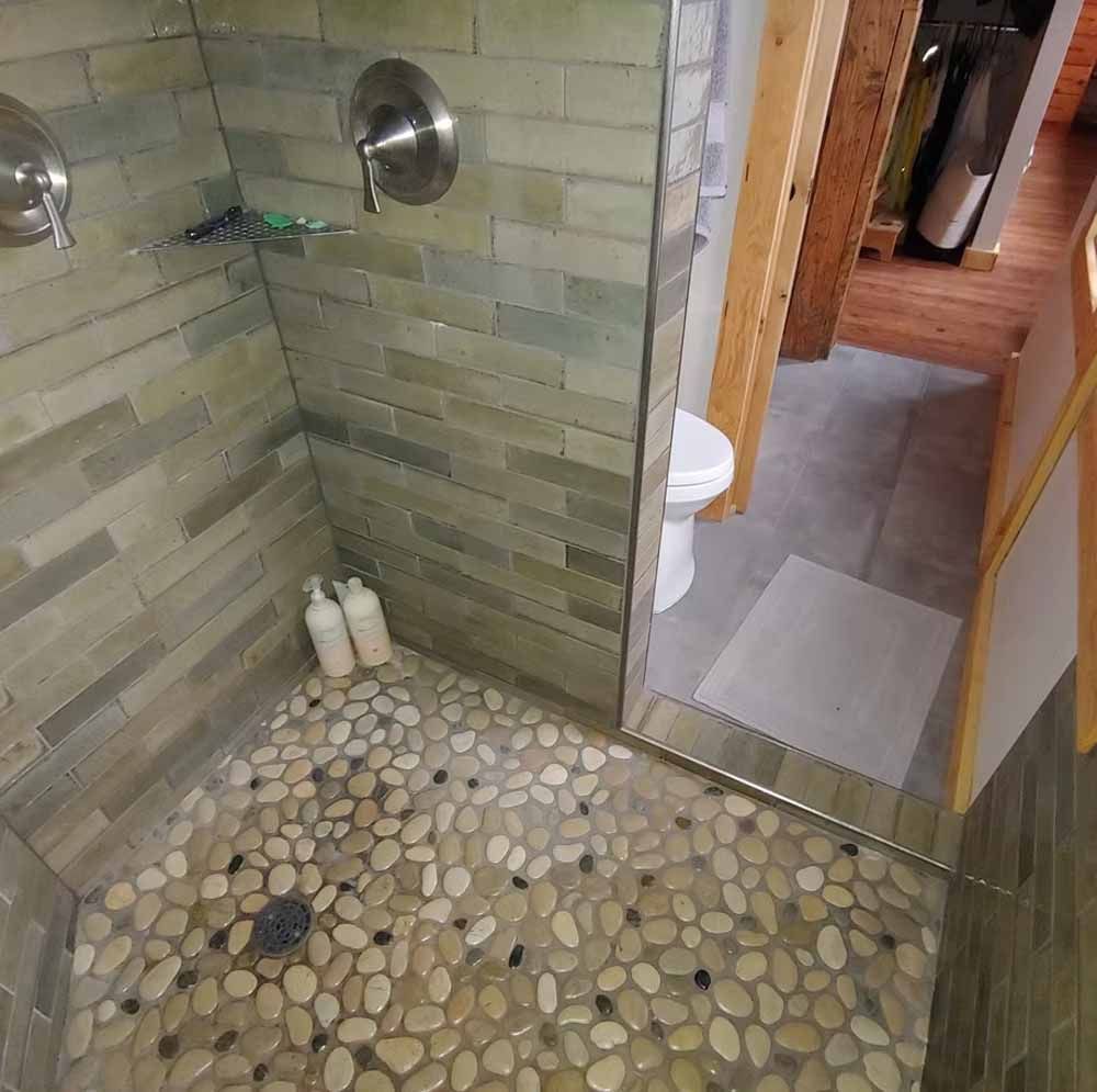 Small tiled shower with pebble floor, green walls, and a view into a bathroom and hallway.