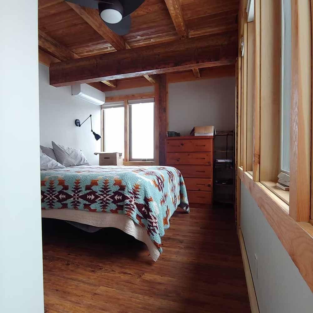Cozy bedroom with wooden ceiling, bed with patterned blanket, dresser, and large window