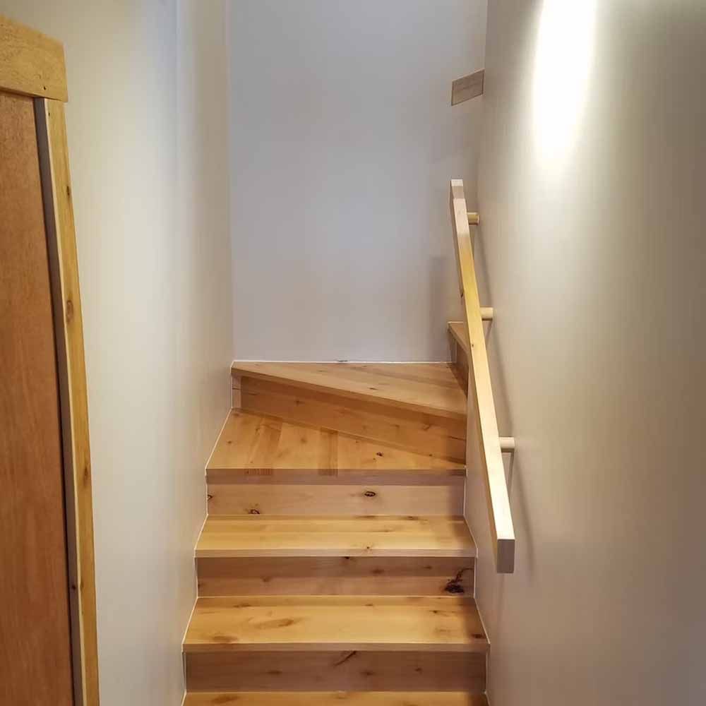 Narrow wooden staircase with a handrail leading up between white walls in a small hallway