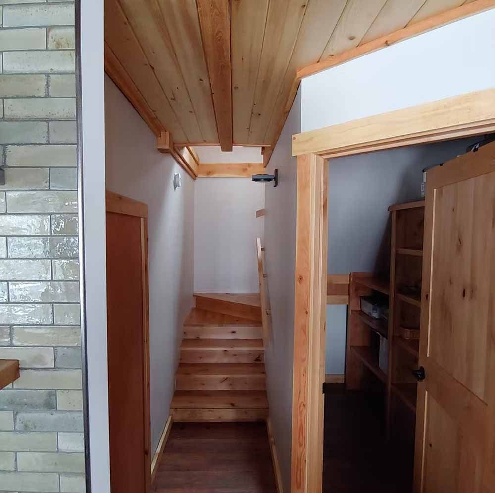 Narrow indoor stairway with wooden steps, white walls, and a sloped wood ceiling beside a hallway.