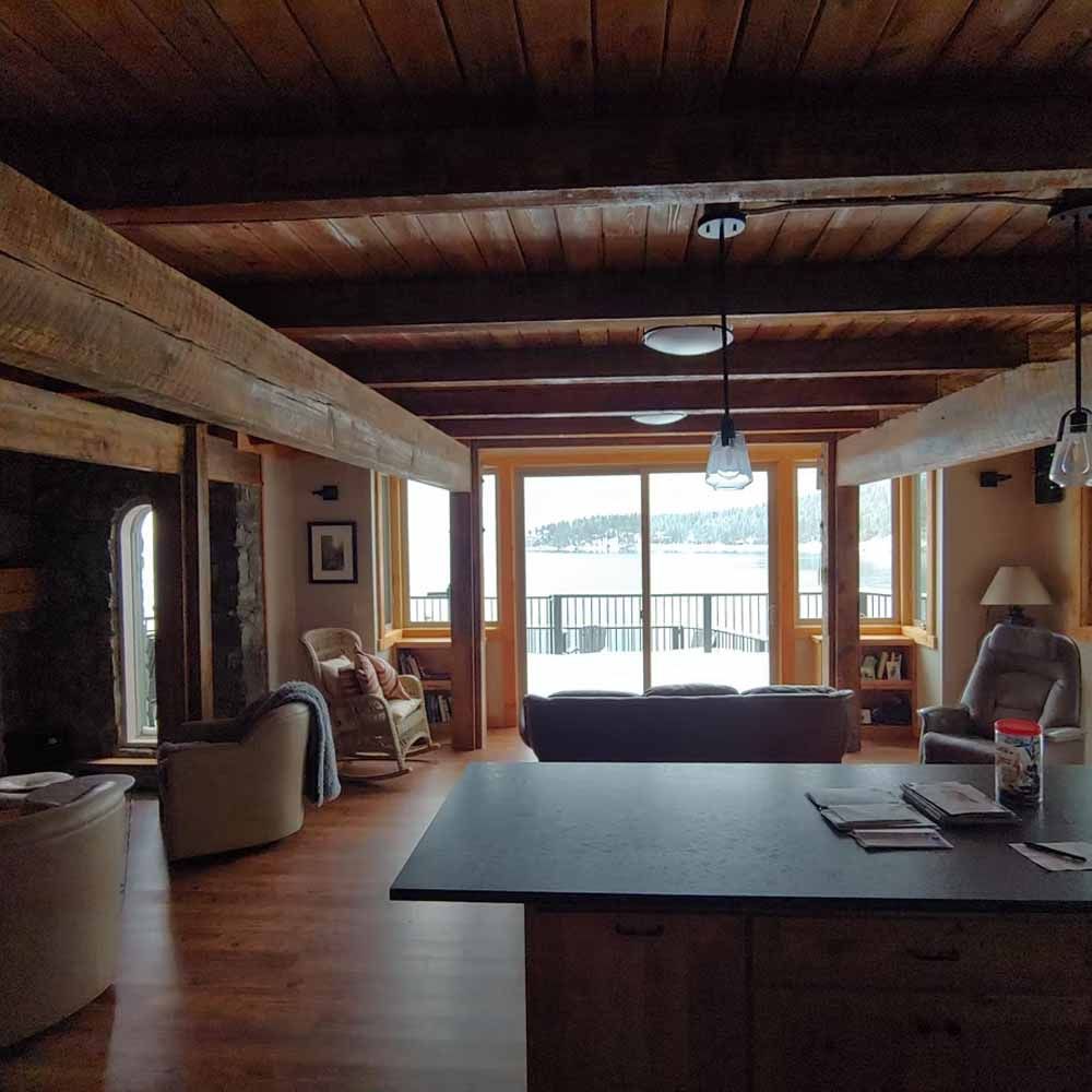 Rustic living room with wood beams, central island, and large windows overlooking a deck and water.