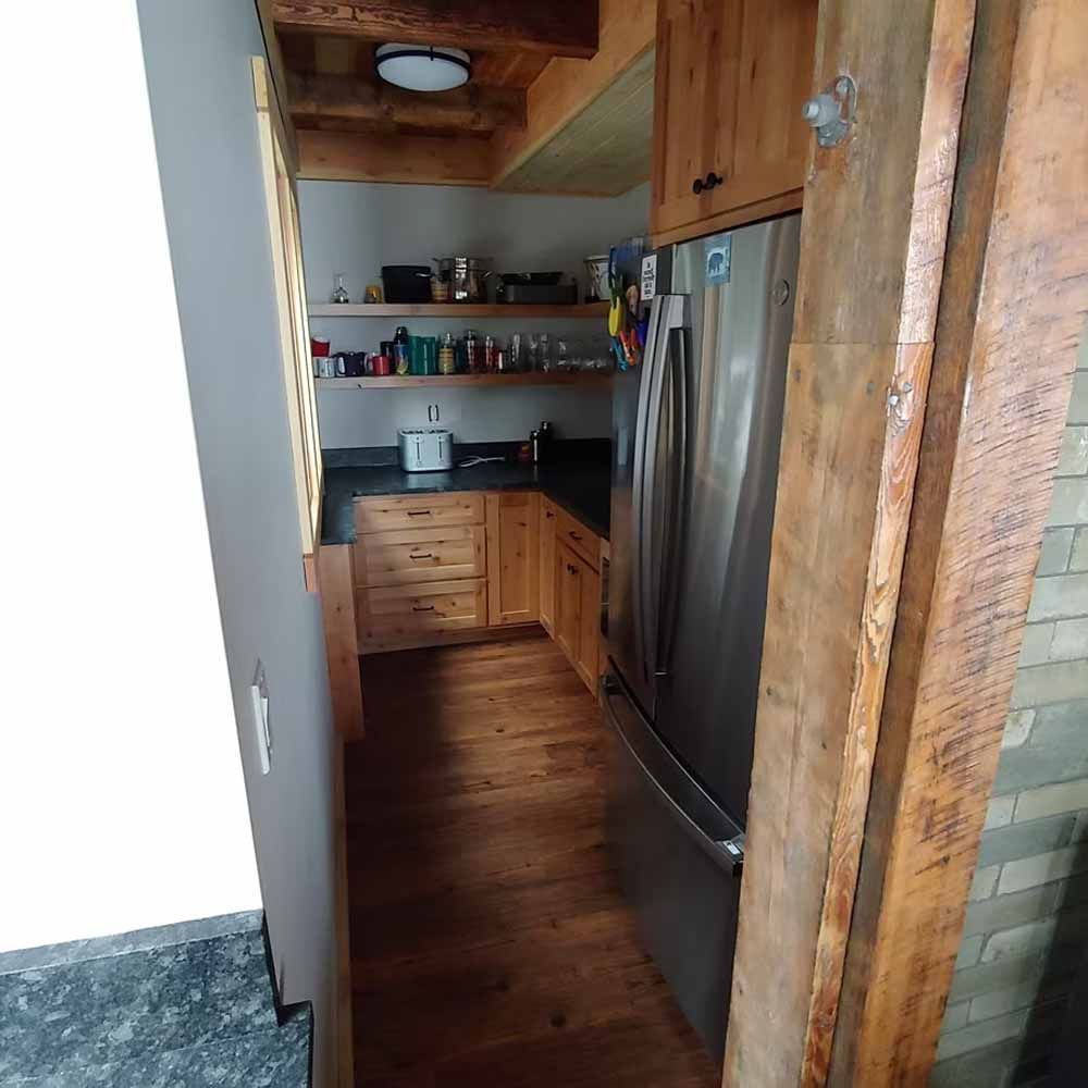 Narrow rustic kitchen with wooden cabinets, stainless steel fridge, shelves, and wood-paneled ceiling.