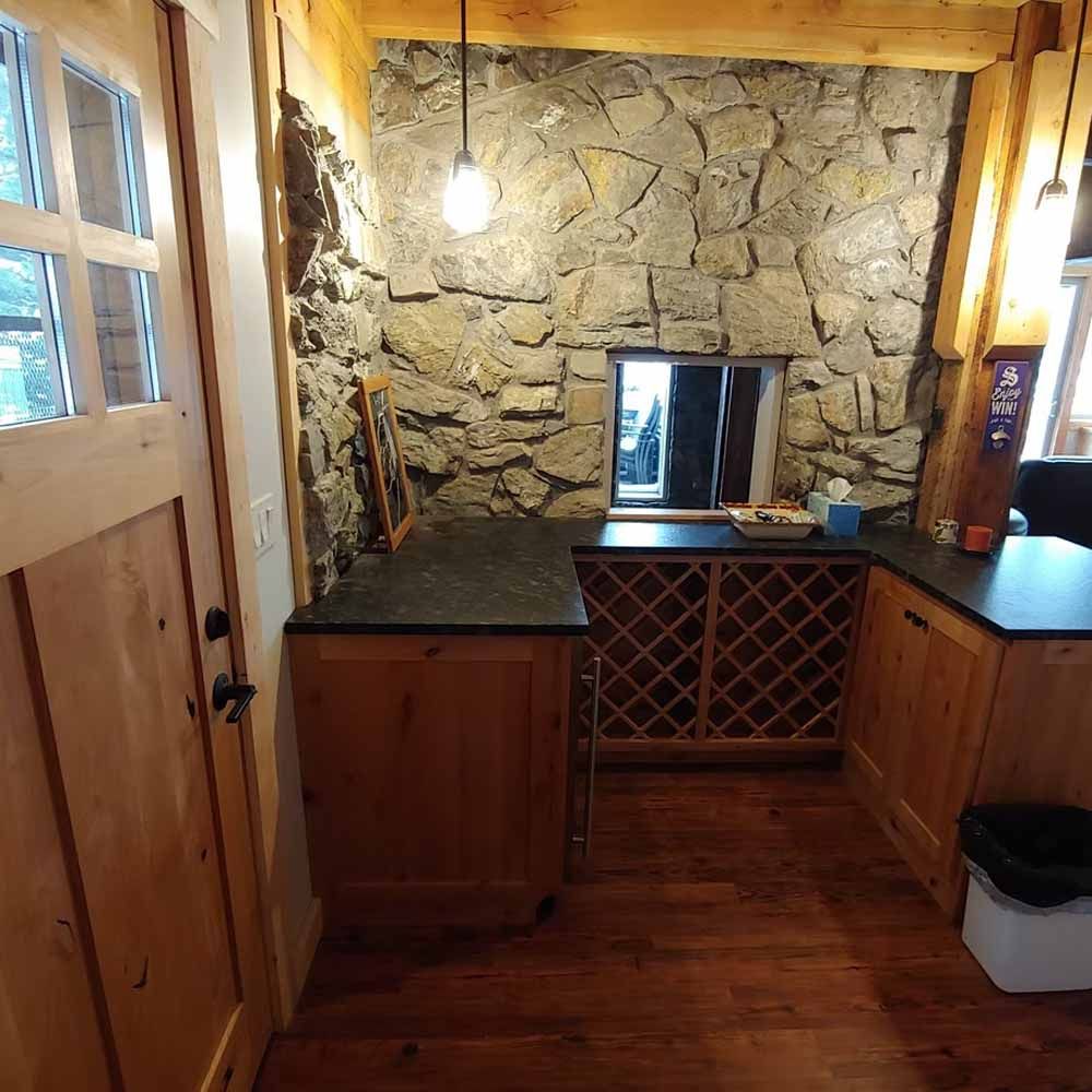 Small rustic kitchenette with stone wall, wood cabinets, granite counters, and a window under a hanging bulb