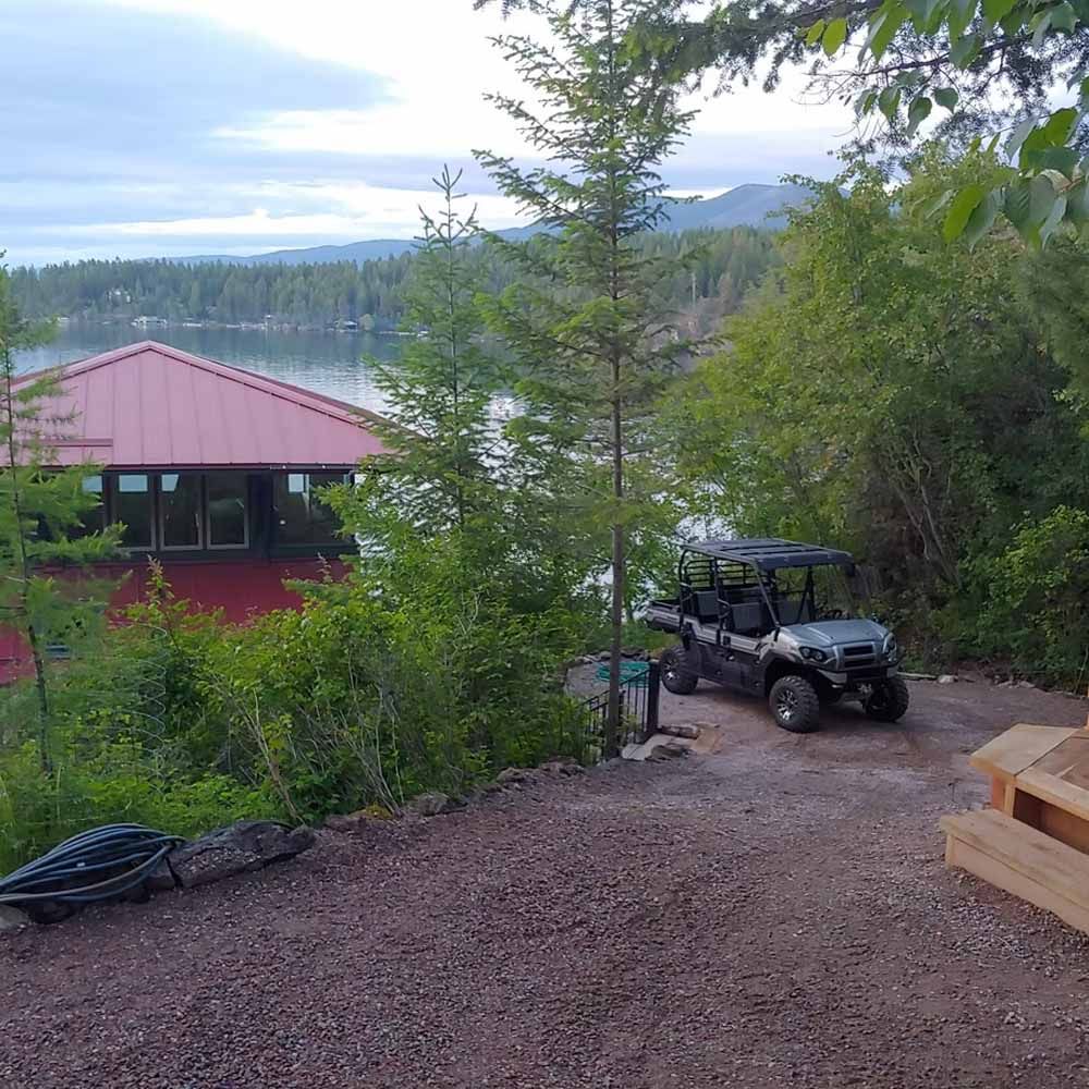 Off-road vehicle parked by a gravel driveway overlooking a lake and forest, with a red-roofed cabin nearby