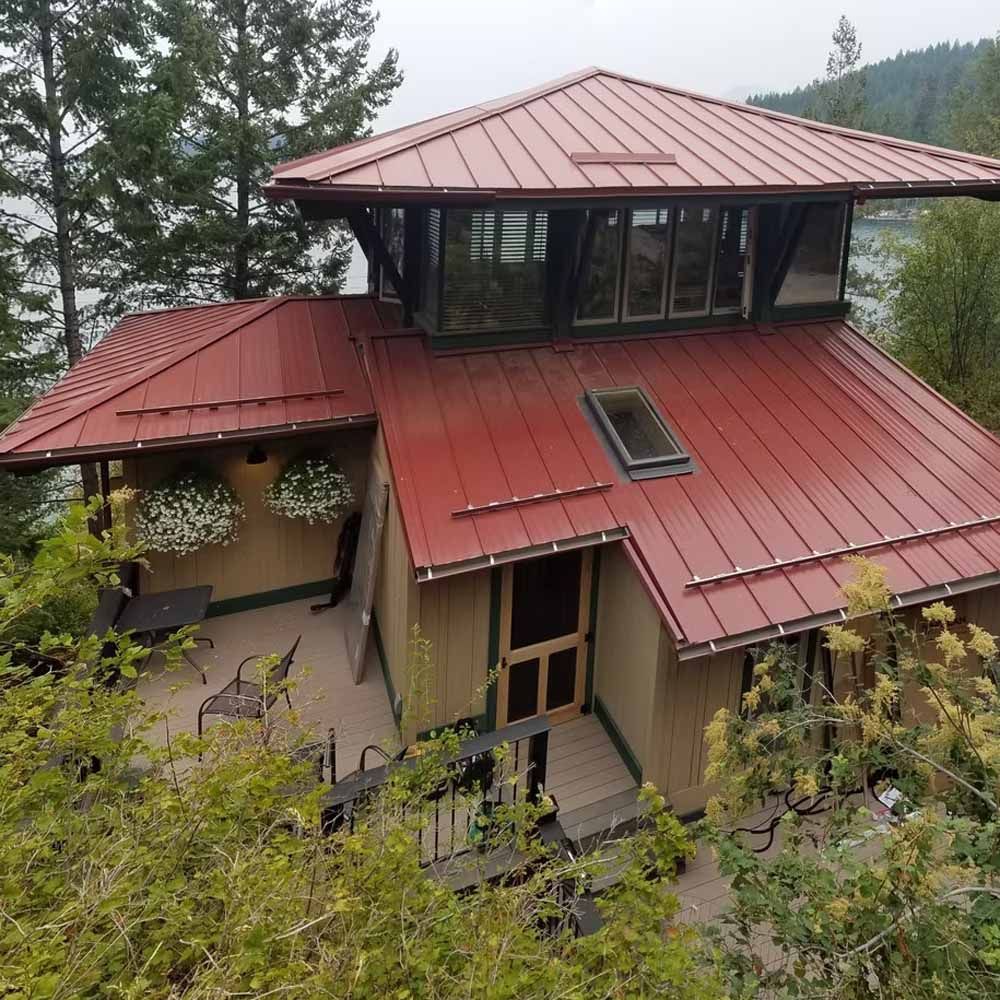 Two-story house with red metal roof and large windows, nestled among trees on a hillside