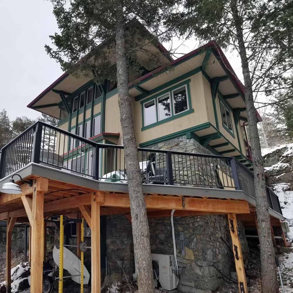 Elevated cabin with green-trimmed windows and black railings among tall trees in a snowy setting