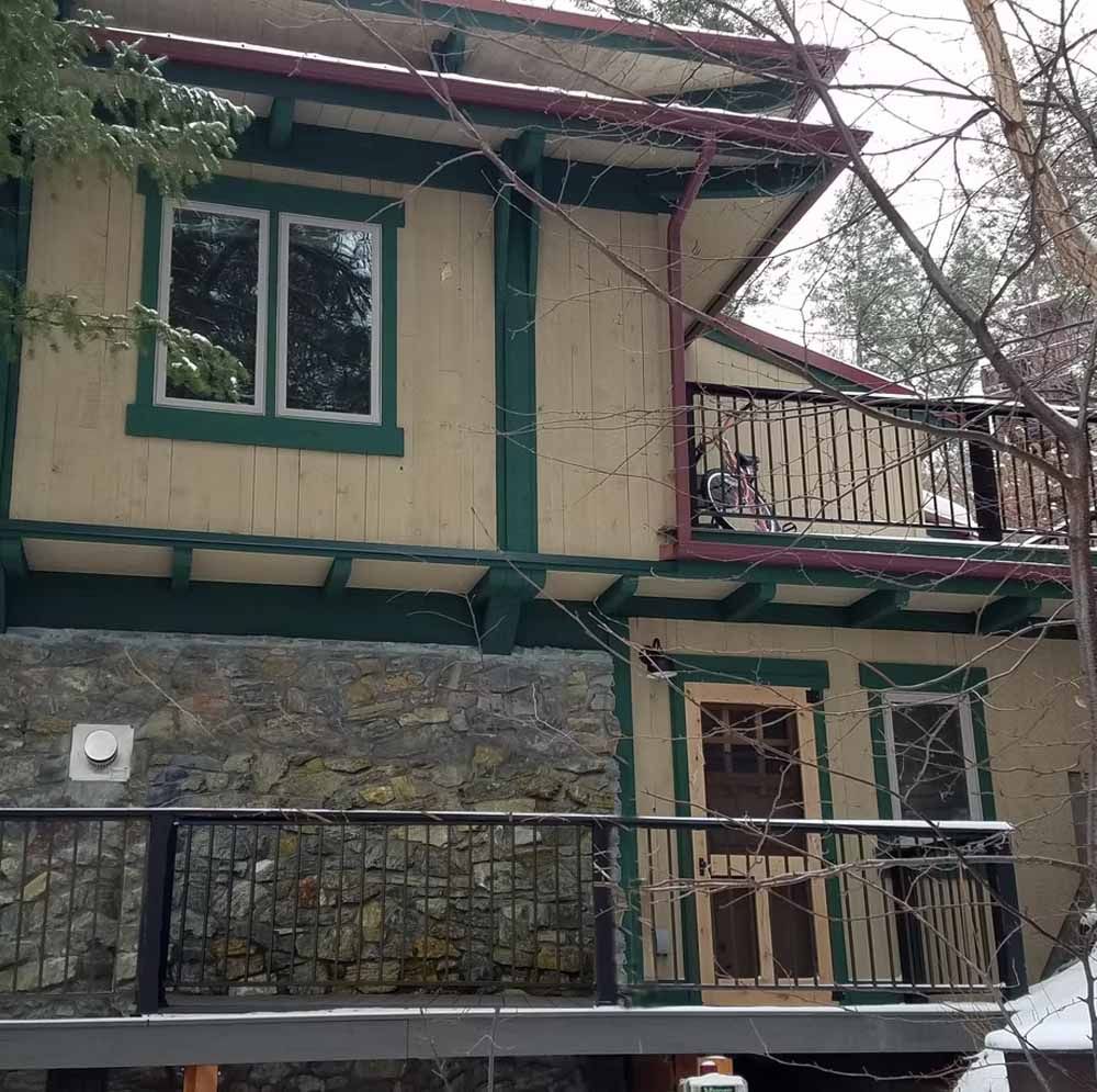 Two-story house with green trim, stone lower walls, and balconies behind leafless tree branches