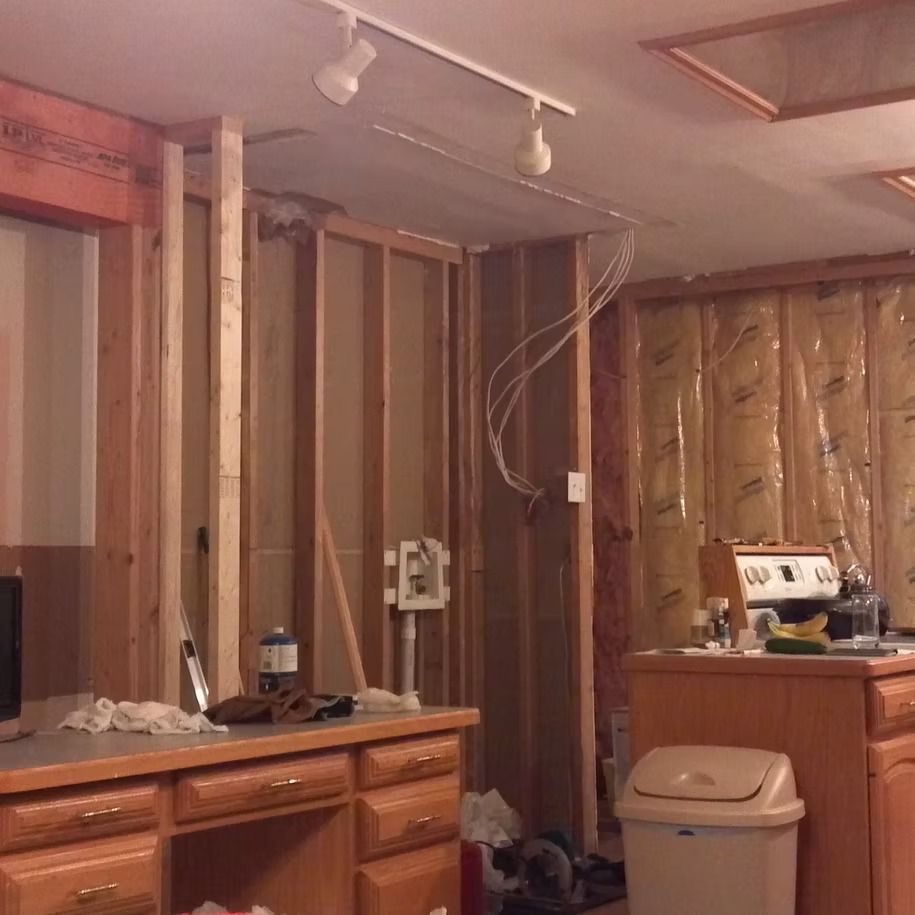 Unfinished kitchen with exposed studs, cabinets, countertop, trash can, and hanging ceiling lights.