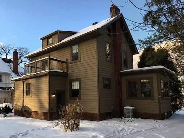 Snow-covered two-story tan house with a small balcony and bare trees in winter.