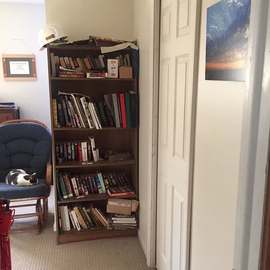 Narrow hallway with a leaning bookcase, white doors, and a blue chair beside a wall picture.
