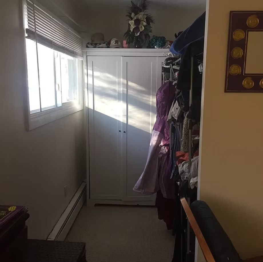 Narrow hallway with sunlight through windows, a white cabinet, hanging clothes, and a framed wall decoration.