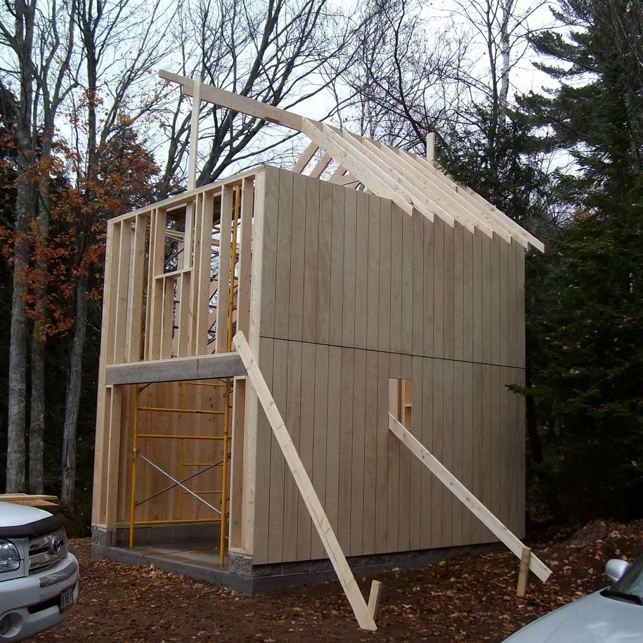 Wood-framed house under construction with plywood siding and roof trusses in a wooded lot