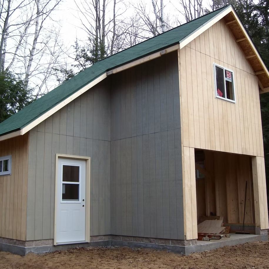 Partially finished beige and gray shed with green roof and white door in a wooded area