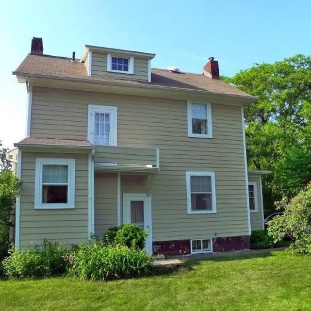 Two-story beige house with white trim and a red brick foundation, surrounded by green lawn and trees