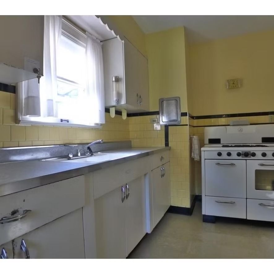 Small yellow kitchen with white cabinets, sink under a window, and a white stove on the right.
