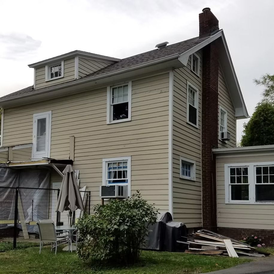 Beige two-story house exterior with white trim, chimney, and a small porch amid yard clutter