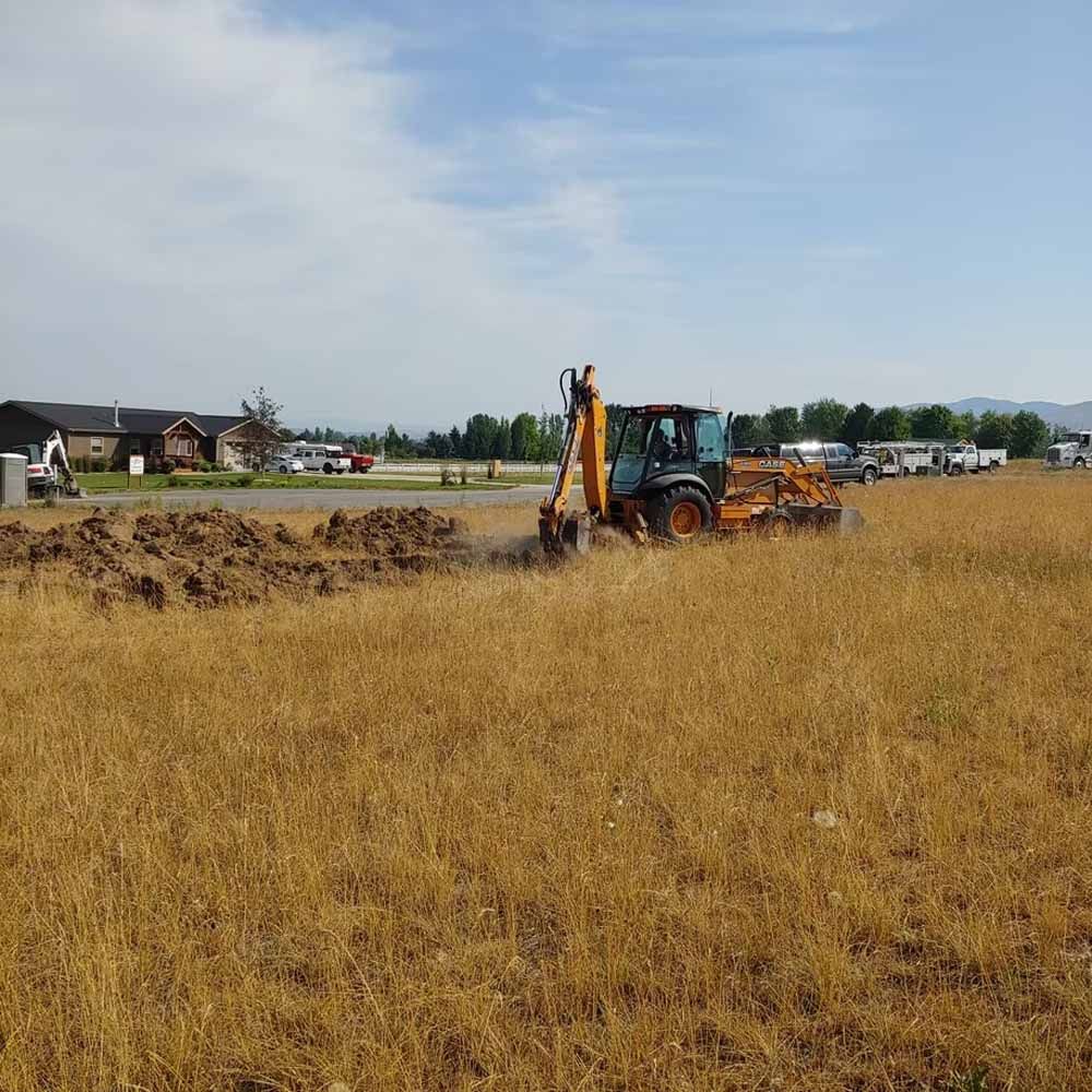 Backhoe digging a trench in a dry field beside a road and houses under a cloudy sky