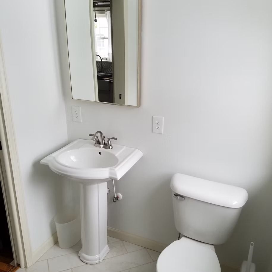 Small white bathroom with pedestal sink, mirror, and toilet against a white wall.