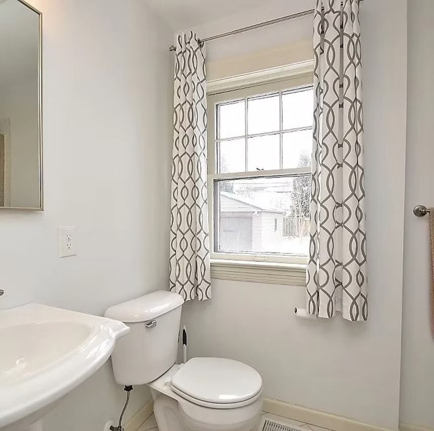Bright bathroom with white toilet, sink, window, and patterned curtains.