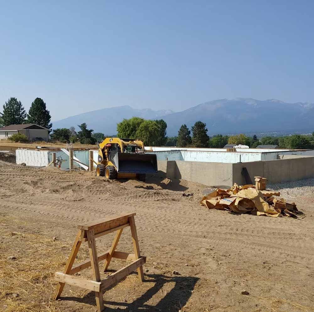 Bulldozer working on a dirt lot beside a white building, with mountains in the background.