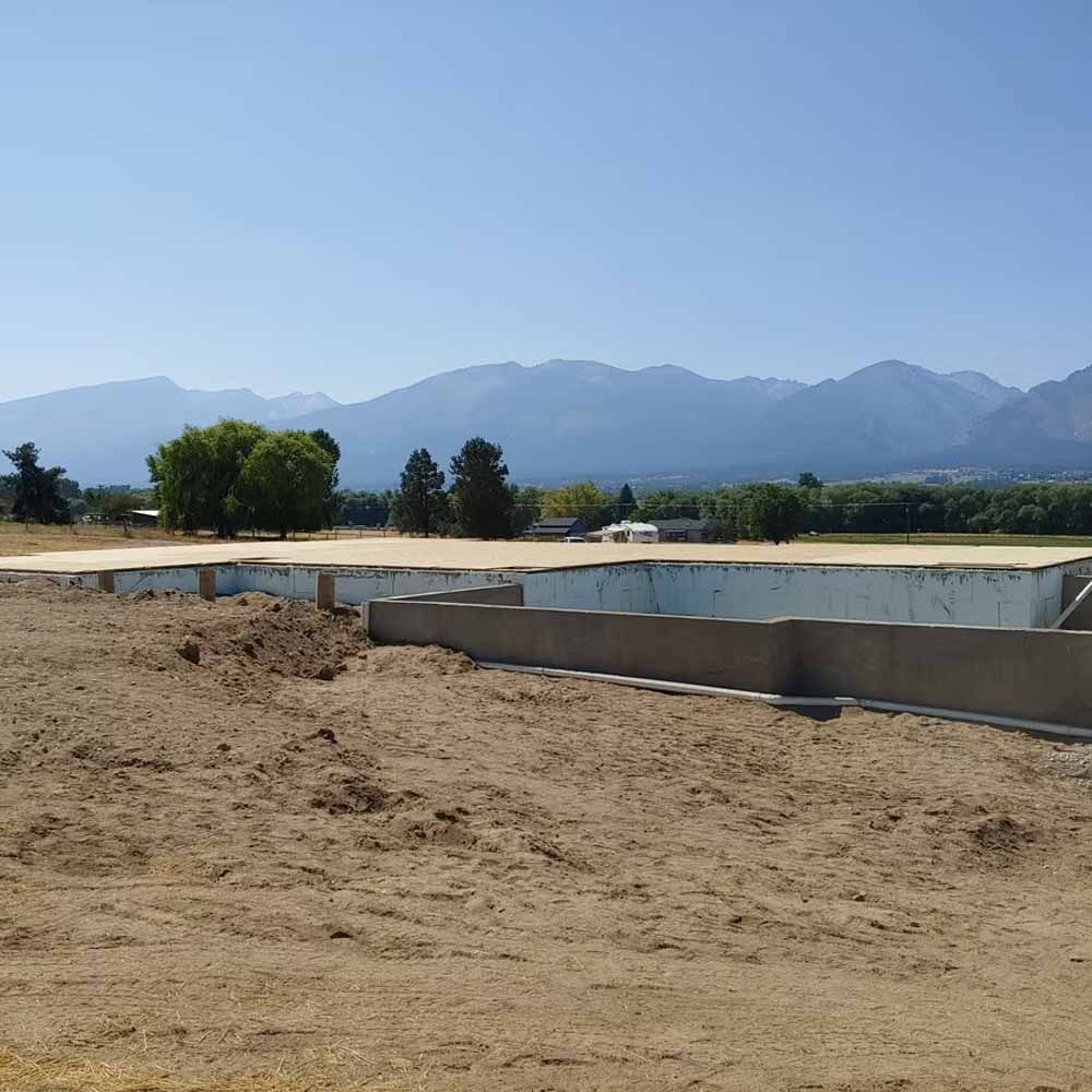 Dry construction site with concrete foundation, sandy ground, trees, and mountains under a clear blue sky
