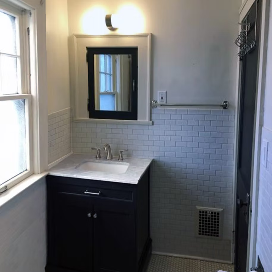 Small bathroom with dark vanity, white tile, mirror, wall light, window, and towel bar