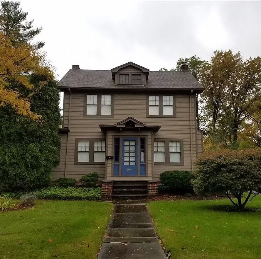Two-story gray house with blue front door, centered walkway, and autumn trees on a cloudy day
