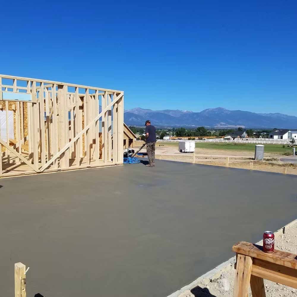 Worker smoothing fresh concrete slab beside a wooden house frame under a clear blue sky.