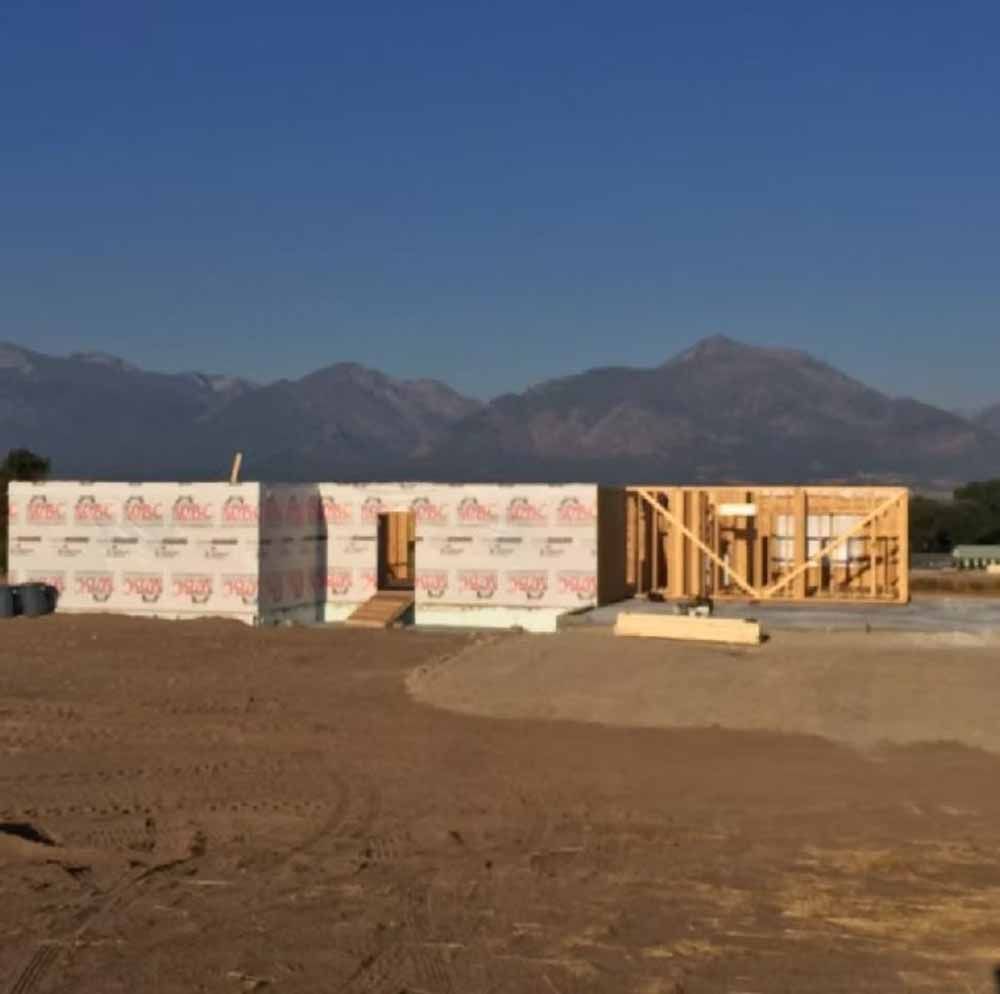 Partially built house framing and wrapped walls on a dirt lot with mountains in the background