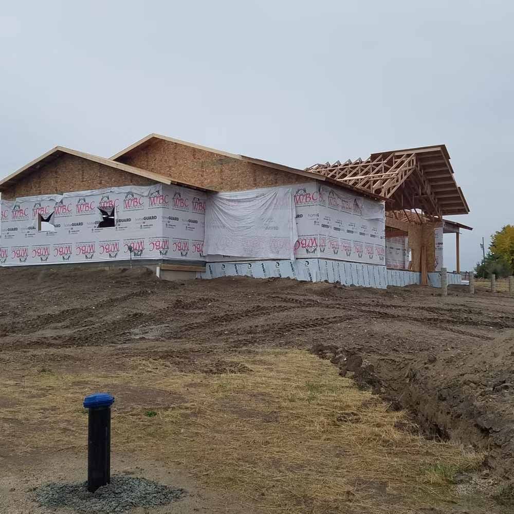 Partially built house with white wrap, wooden roof trusses, and muddy ground in a rural setting