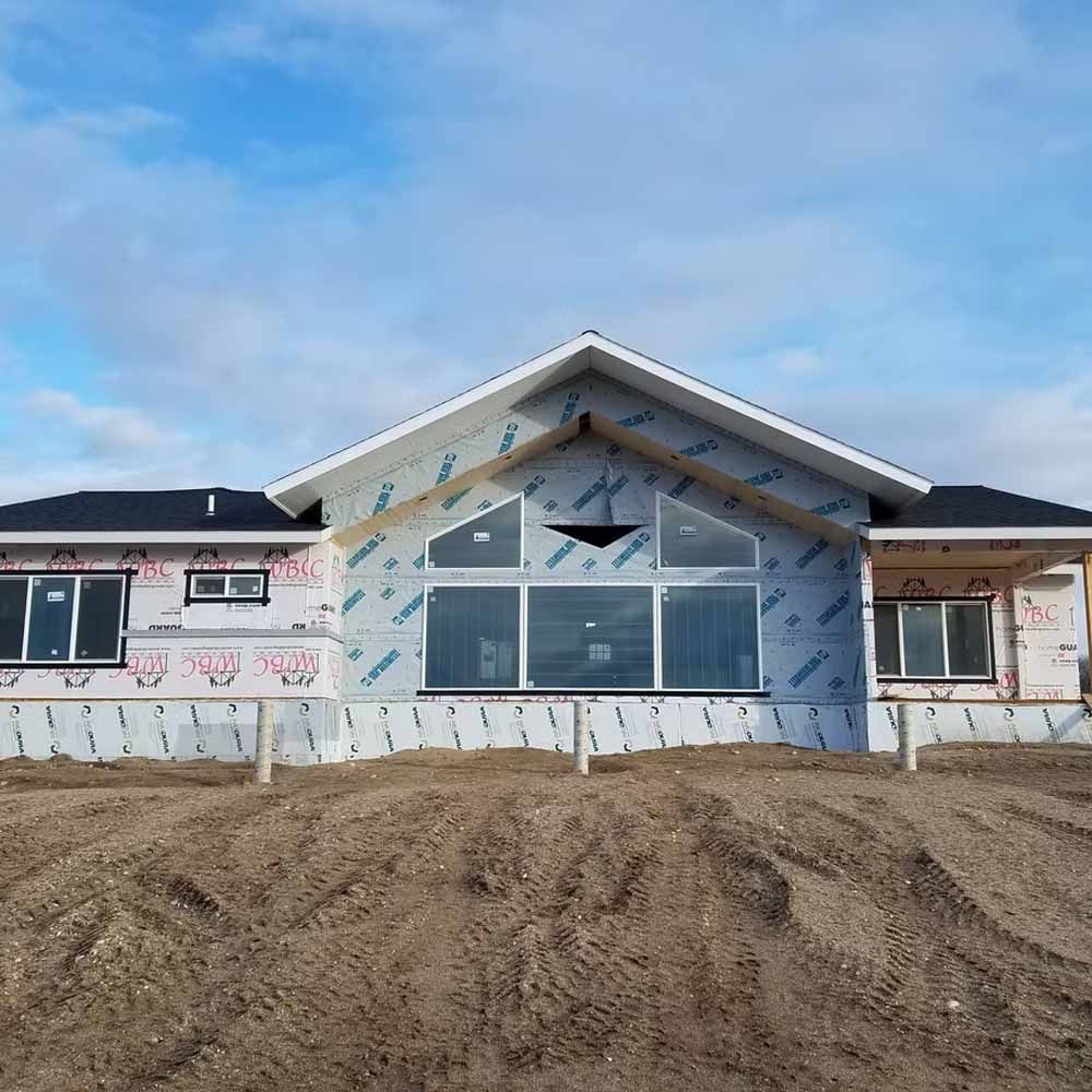 Partially built house with exposed framing, blue wrap, and dirt mound in front under a clear sky
