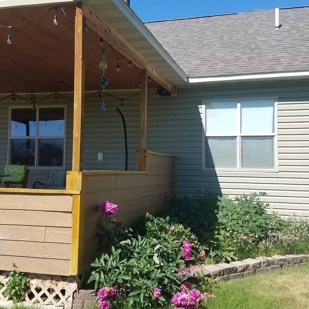 Front porch with wooden railing and hanging wind chime beside a gray house, with pink flowers in the garden.