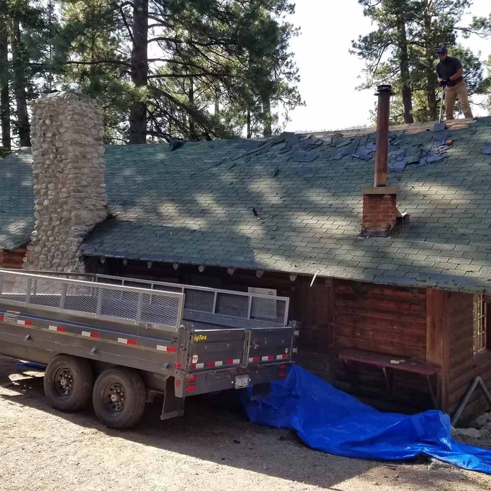 Worker on a cabin roof beside a chimney, with a flatbed trailer and blue tarp below