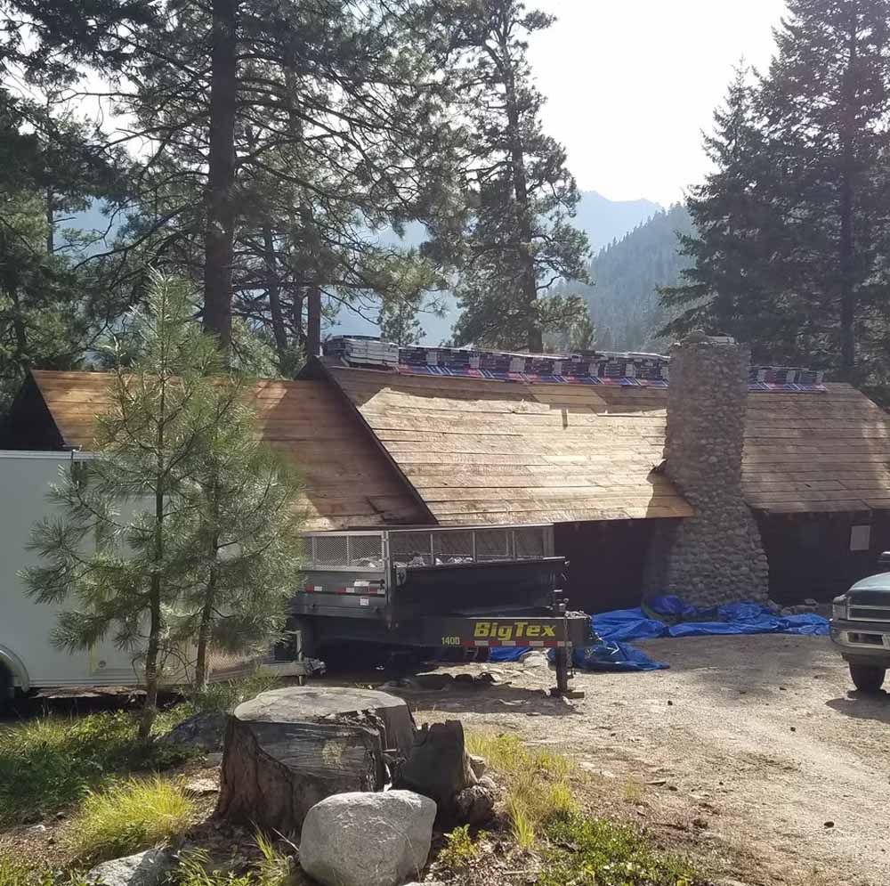 Rural cabin with wooden roofs among pine trees, with rocks and a parked truck in the foreground.