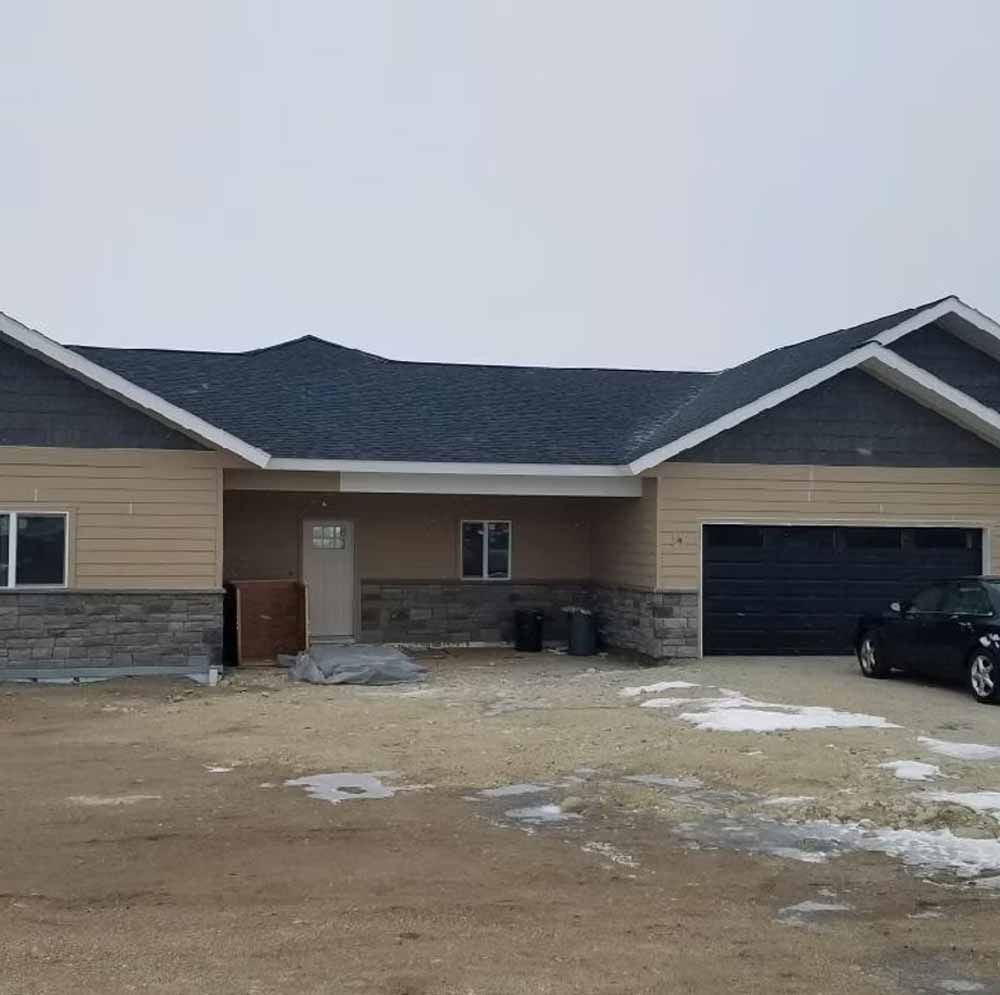 Single-story beige house with dark roof and attached garage on a snowy, muddy lot