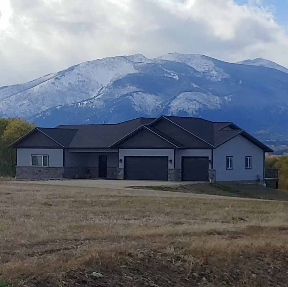 Single-story gray house with a dark roof in front of a snow-dusted mountain under cloudy sky