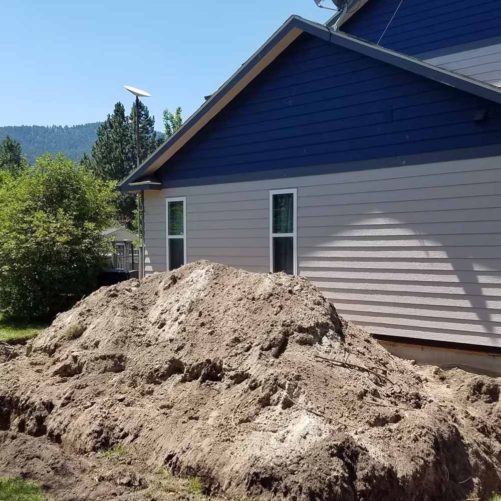 Large pile of dirt beside a blue house with beige siding on a sunny day