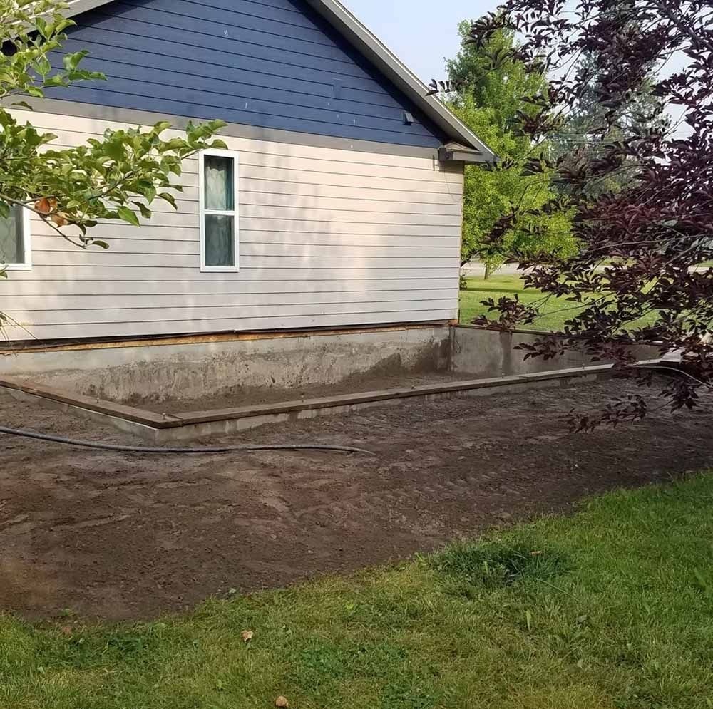 House side with exposed foundation and dirt yard beside grass and trees