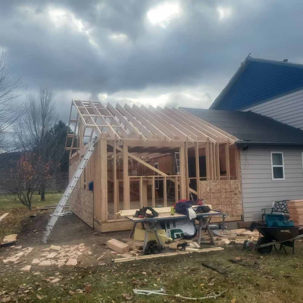 Partially framed house addition under construction beside a gray house, with tools and debris in front