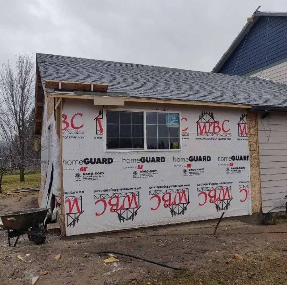 Small shed under construction with white house wrap and a gray shingled roof outdoors.