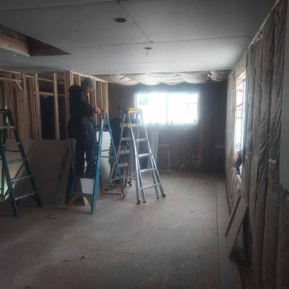Person on a ladder in an unfinished room with exposed framing and bright windows.