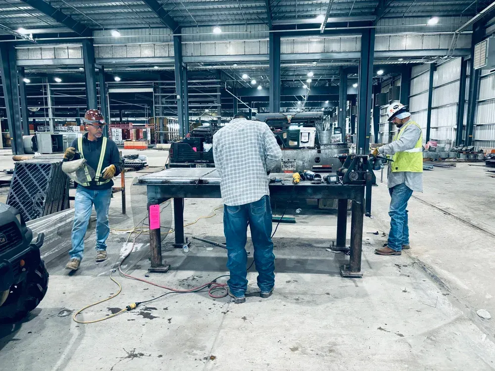 Three construction workers in a workshop; one welding, two working at a metal table, wearing safety gear.