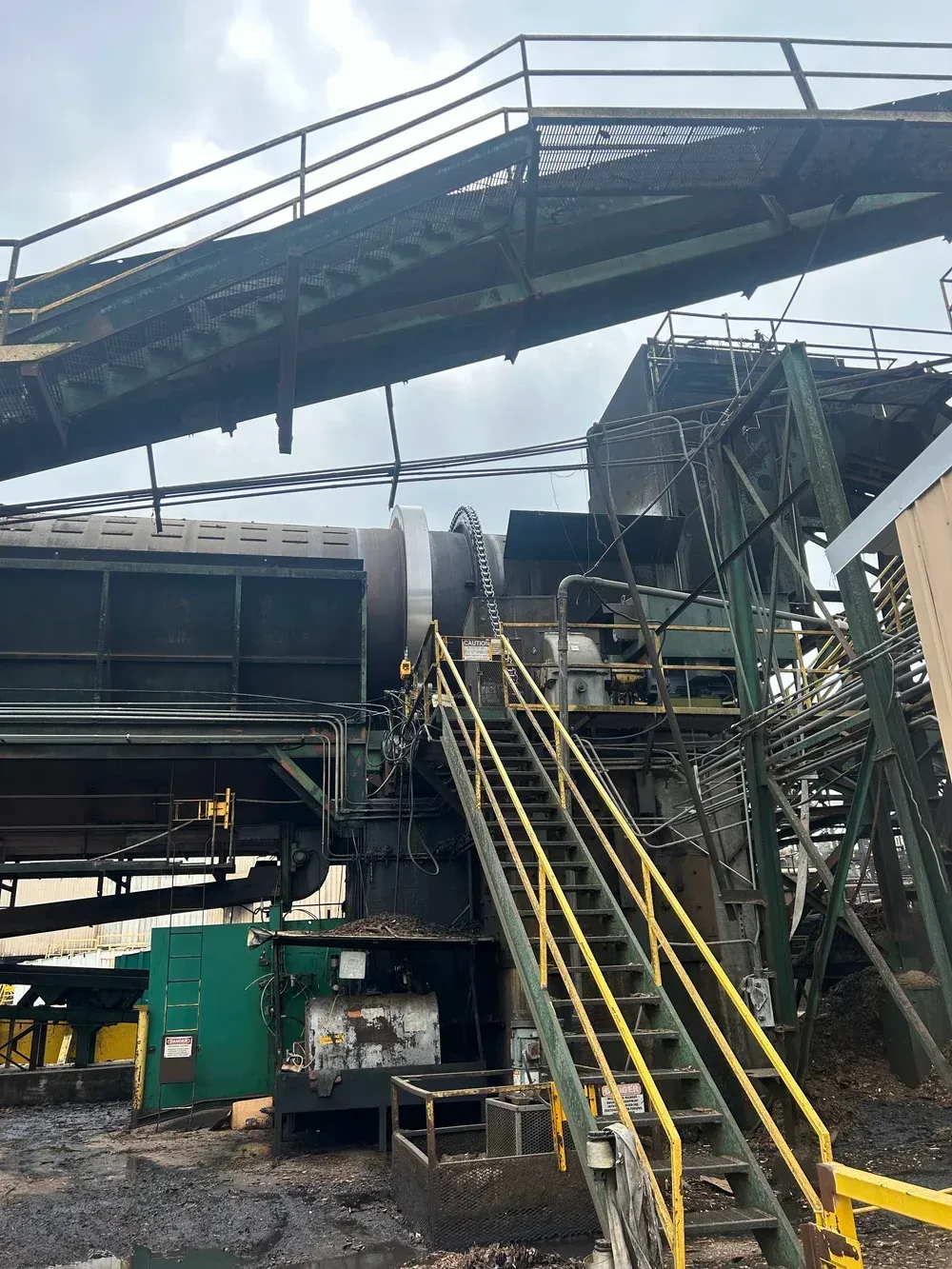 Industrial machinery with stairs, catwalks, and a person. Black residue suggests fire damage, cloudy sky.