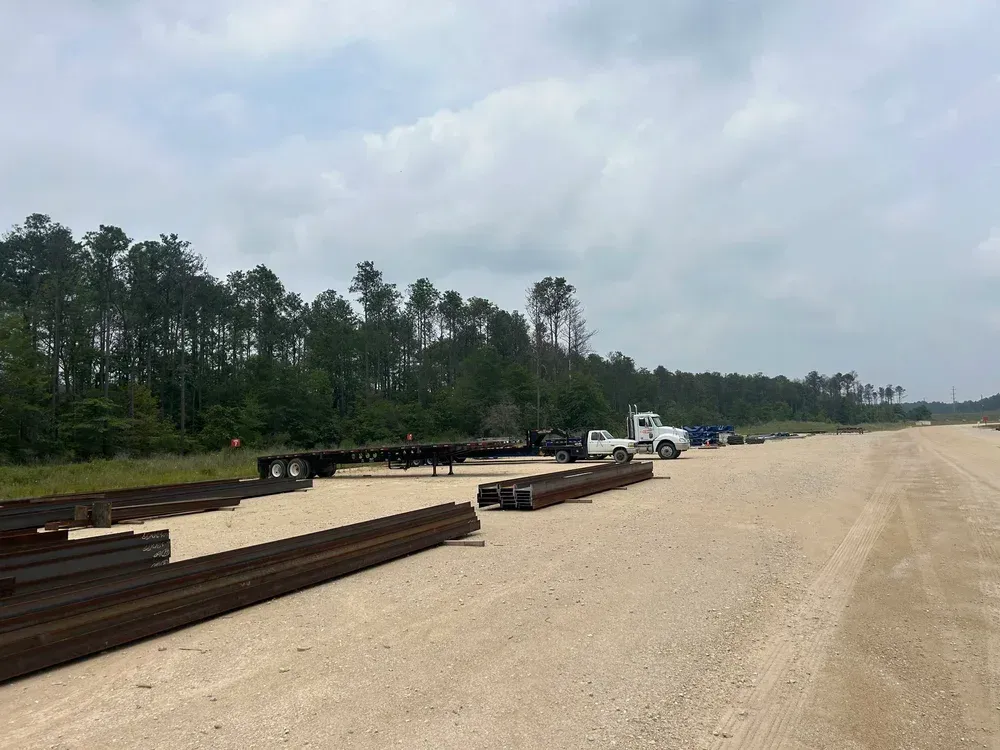Construction site with trucks, trailers, and steel beams on a gravel road, forest in the background.