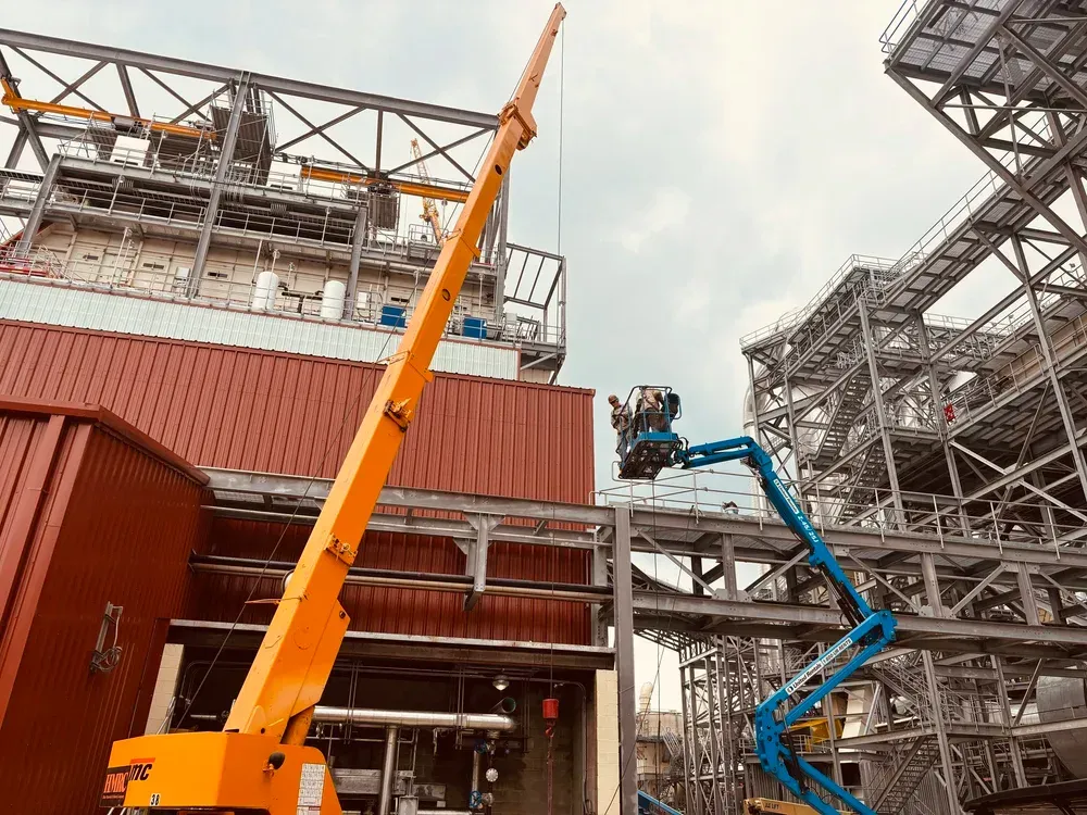 Two aerial lifts at a construction site; orange lift extended near a red building and a blue lift working on metal framework.