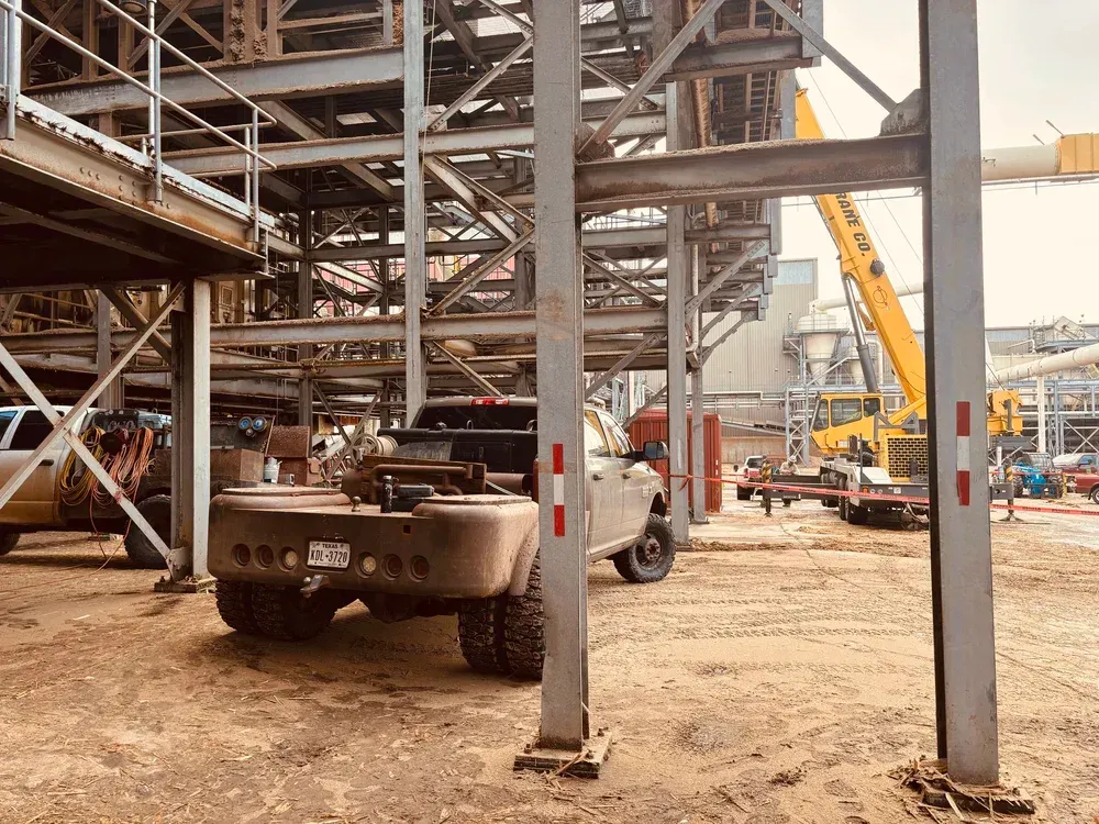Construction site with a truck parked under a metal framework, and a yellow crane in the background.