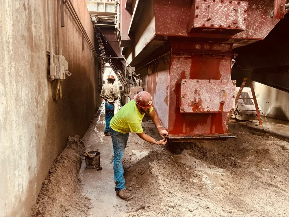 Two workers near industrial machinery; one inspects it, other in background. Red machinery, brown dirt, narrow space.