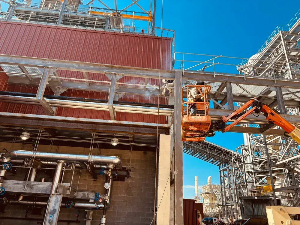 Worker in an orange lift operating near red structure and industrial scaffolding under a blue sky.