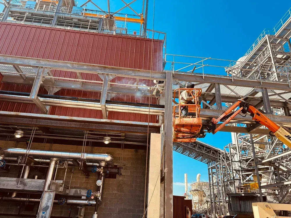 Construction worker on lift welding steel beams; industrial site, blue sky.